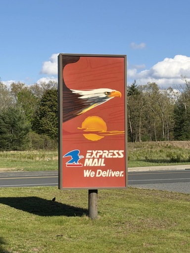 A photo outside with a tall sign in the foreground. The sign is an advertisement for the Express Mail Service by the United States Postal Service.